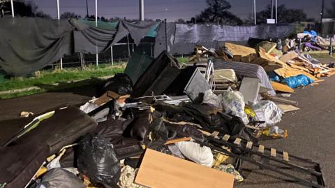 A large amount of rubbish including mattresses, black bin bags and wooden boards dumped in a car park by a football ground.