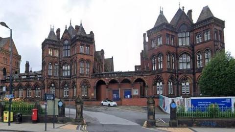 Leeds General Infirmary is pictured. It is a Gothic-style red brick building with several towers. It features tall, narrow windows and arches. A white car is parked in front of the hospital.