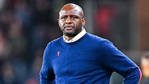Patrick Vieria, wearing a white shirt and a blue jumper, stands with his hands on his hips