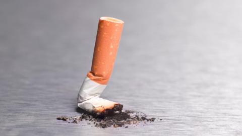 A cigarette has been put out and crushed on a grey table. There is black ash surrounding it, and it is crumpled.