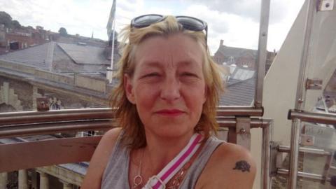 Dawn Sturgess sitting in a glass ferris wheel cabin with silver bars. She is wearing a grey vest, black sunglasses on her hand, a circular necklace and a white and pink striped bag strap across her chest. She has shoulder length blonde hair and is smiling at the camera, slightly squinting from the sun.