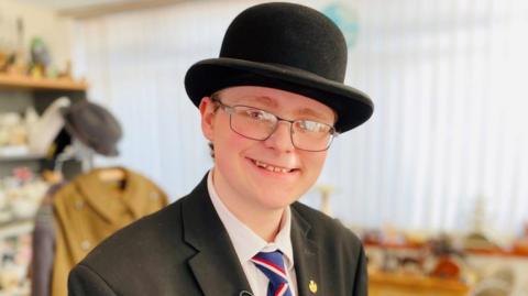 Boy, 16, smiling, wearing glasses, a black bowler hat, black jacket, white shirt, red white and blue striped tie, with antiques in the background.