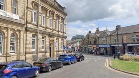 Four cars parked along a road in Brighouse, opposite a parade of shops.