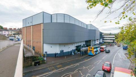 General view of Merrywalks transport hub, which has blue panels and white wall. Buses and cars are traveling past on road.