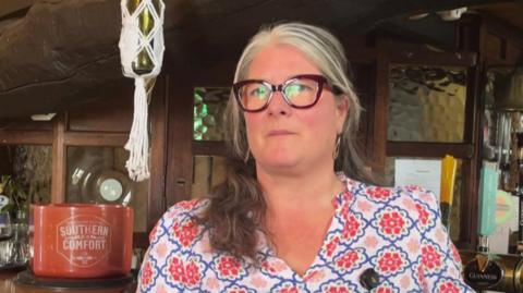 Lou Tresidder is resting her arm on the bar. She has long white and grey hair and is wearing glasses and a brightly patterned blouse.
