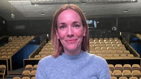 Actress Laura Main smiles at the camera from the stage in Aberdeen Arts Centre, the seats of the auditorium behind her. She wears a pale lilac mohair jumper and her bobbed hair is tucked behind her ears