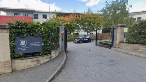 The entrance to a school, as marked by an open set of black gates. A sign with the school's name is on the fence to the left of the gates. A taxi is emerging from the other side.