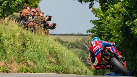 Dean Harrison in red and blue leathers riding away on the TT course. He is leaning over towards a grass embankment where a group of spectators are holding up mobile phones filming him.