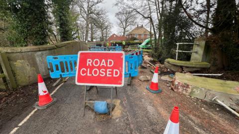A general view picture of the bridge looking towards homes in Swallowfield with a red sign saying "road closed" in front of the bridge, which has been damaged. Part of the smashed bridge is pictured on the far right side of the picture with metal railings sticking from its top.