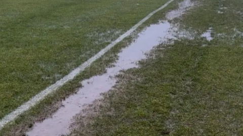 A close up of a long puddle in waterlogged ground on a grassy football pitch. The water runs parallel to a painted white line on the pitch.