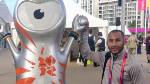 Demetrios Bradshaw, who is wearing a grey tracksuit, poses for a photo next to a statue of the large, silver Olympic mascot Wenlock.