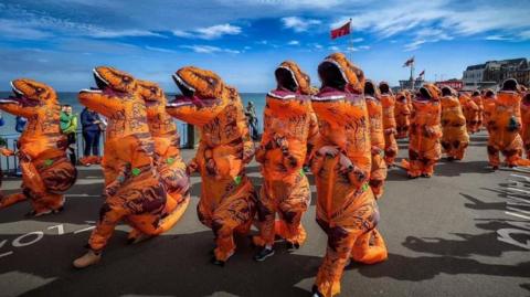 A crowd of runners in inflatable dinosaur costumes run along a promenade next to the sea.