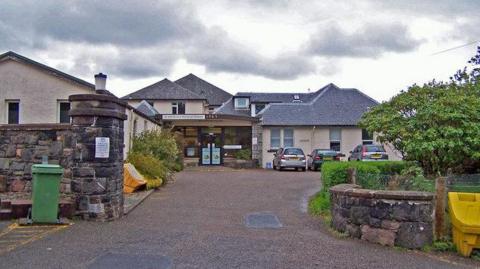 The hospital is made up of a number of low buildings with peaked, slate roofs. Three cars are parked near the glass entrance. 