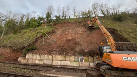 A large brown earth embankment next to a railway line that has suffered a landslip. There are layers of concrete blocks at the bottom and a large orange digger with a cherry picker is reaching up to the top of the embankment.
