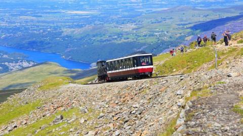 Train carriage seen from the side of mountain as it climbs up, with lake visible in the distance and rocks and grass alongside the tracks. The train is made up of one carriage painted red, white and black and hikers are seen walking on the mountain a little further up