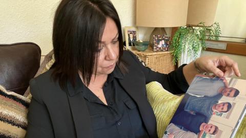 A woman in a black jacket and shirt sits on a sofa looking at a photo of three young men.