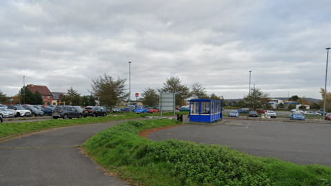 A large car park filled with cars. There is a curved road area for buses, and a blue bus shelter.