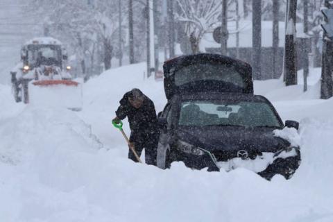 A man digs a black car out from thick snow while a tractor ploughs snow behind him