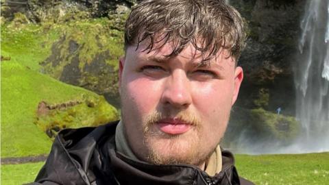 Close up selfie-style head shot of a man standing in front of a lush green backdrop featuring a hill side and a waterfall. He has brown hair - combed forward with a wet look - and a wispy blonde moustache and goatee beard. He is wearing a black hooded jacket.