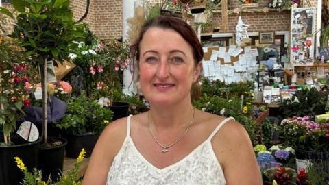 There is a woman who is wearing a spaghetti lace white t-shirt and a pendant. She has dark hair tied up. Behind her is lots of colourful flowers and plants, plus a big noteboard over her left shoulder.