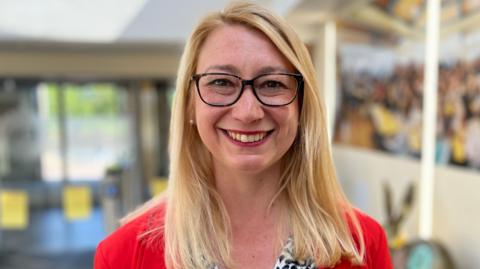 Jess Asato, wearing a black and white patterned blouse, with a red blazer. She has blonde hair and is looking into the camera and smiling and has black rimmed glasses on. 