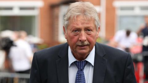 Sammy Wilson looking at the camera with a neutral expression. He is wearing a suit and tie. There are people and a building in the background very out of focus.