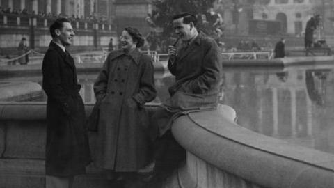 Three people smile and talk while sitting on the edge of a fountain.