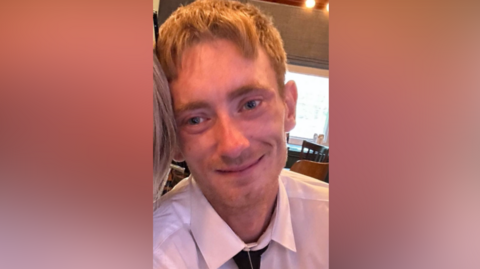 Conor Kinlan is a man with blond hair, wearing a white, collared shirt and a black tie. He is looking directly at the camera and is smiling.