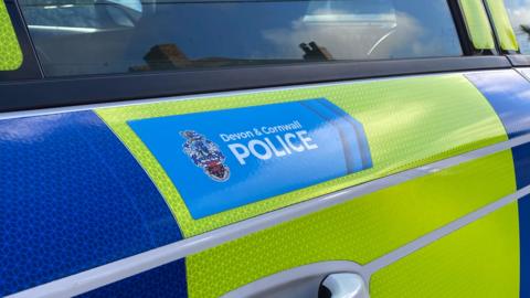 The side of a yellow and blue police vehicle. There is a blue section with the words 'Devon and Cornwall Police' inside of it. The door handle is below and a window above the section.