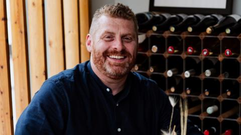 A man with short ginger hair and a ginger beard, and wearing a blue buttoned shirt, smiles as he sits in front of a large wine rack full of bottles of wine.