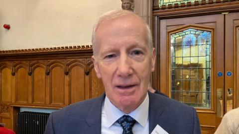 Shows a man with receding grey hair, a blue jack, light-coloured shirt and blue floral tie in the Guildhall in Derry. 