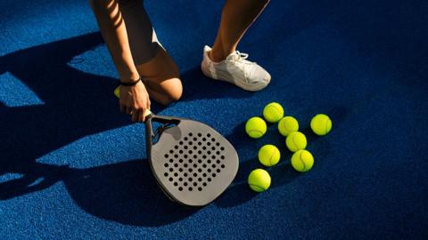 Close up of a player holding a padel racquets by a blue padel court on which are a number of yellow tennis balls