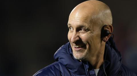 Alan Dickens wearing an ear piece in his left ear, looking to his right and with a smile on his face, ahead of the recent match at Sale  