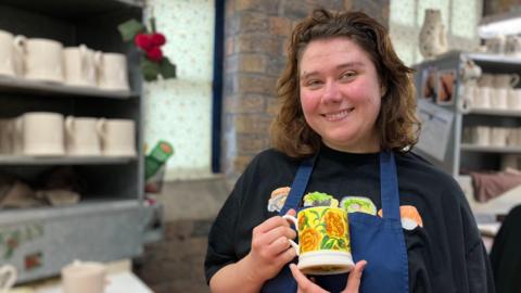 A woman who is smiling directly at the camera whilst holding a mug which has a lithograph stencil placed on it. She is wearing a blue apron with a black t-shirt underneath that has pieces of sushi printed on it. She has brown hair and an eyebrow piercing over her right eye.