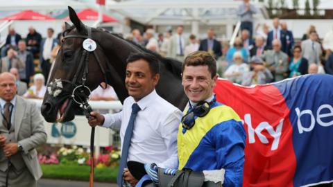 Oisin Murphy wearing a blue and yellow top and another man wearing a white shirt and blue tie, holding the reins of a dark brown horse. There's a crowd of people behind them.