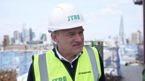 Ed Davey being interviewed. He is wearing a hard hat and a yellow high vis tabard. The London skyline is in the background. 