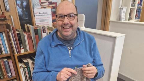 A bald man wearing glasses and a blue jumper knitting in front of a bookshelf 