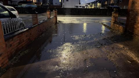 A large pool of water reflects streetlights and spans a domestic dirt-track lane between two brick walls 