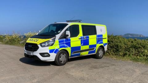 A white, yellow and blue Ford police van parked up in a coastal car park in Guernsey. The Guernsey Police emblem is on the side of the van which is parked in front of a hedge.