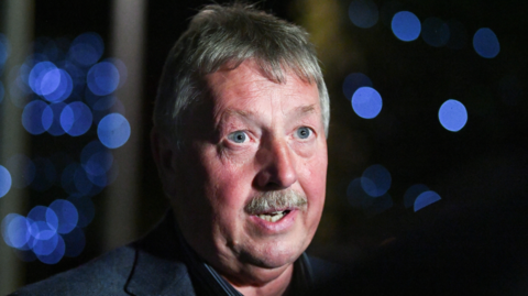 Headshot of Sammy Wilson speaking, looking slightly off camera. In the background some lights that are out of focus.