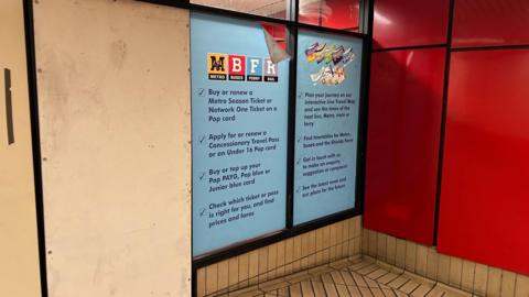 The closed ticket office. The door has been sealed up with plyboard. Posters, advertising the services previously offered at the ticket office, remain on the glass windows but are pealing due to their age.