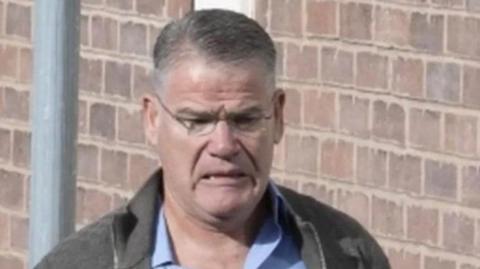 A man in a jacket and blue shirt is wearing glasses. A brick wall is behind him.
