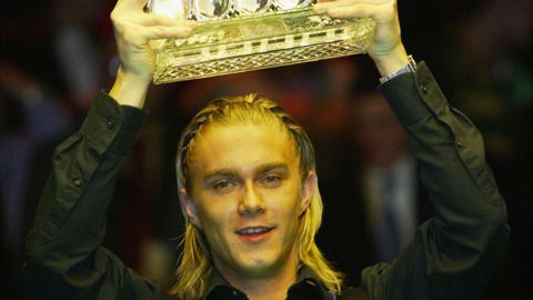 The snooker player Paul Hunter, lifting a trophy aloft out of shot. His blond hair is braided and he is wearing a black shirt.