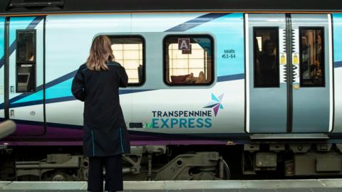 A woman stands on a rail platform at station in front of a green-liveried TransPennine Express train, with passengers seen on board.