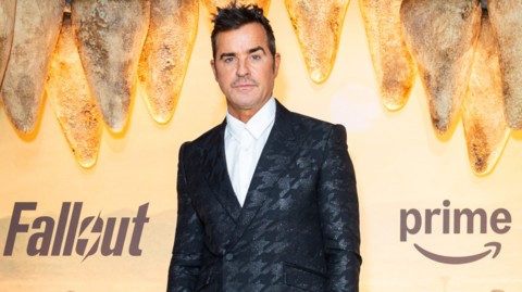 Justin Theroux is standing in front of an orange background, with the words Fallout and Prime on it. He is wearing a black jacket with a white shirt, and he has black, spiky hair. 