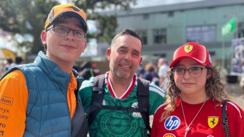 A family of Formula 1 fans stand together for a photo. Guillermo Jr is in a papaya McLaren jersey and gilet. Guillermo Sr is in a Mexico national football team jersey and Rebecca is in a red Ferrari baseball cap and jersey.