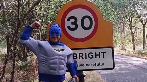 A man dressed a Batman standing next to a road sign.
