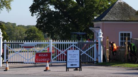 A white gate, Cranbourne Gate, in Windsor Great Park, with two workmen stood to the right of it. One is wearing an orange jacket and the other a green jacket. There is a red "road closed" sign in front of the gate and a sign to its right which says: "This gate is closed, please use Rangers Gate".