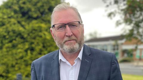 Doug Beattie wearing a blue suit jacket with a white open neck shirt 