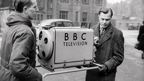 Two men carrying a large camera labelled '91ȱ Television'.